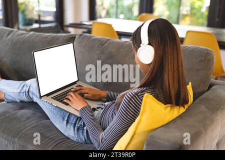 Birassisches Teenager-Mädchen, das Kopfhörer trägt und ein Notebook mit Kopierbereich benutzt. Das Konzept „Zeit zu Hause verbringen“. Stockfoto