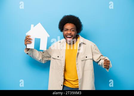 Überraschter afro-amerikanischer Mann, der ein Haus in einem isolierten hellblauen Studio-Hintergrund hält Stockfoto