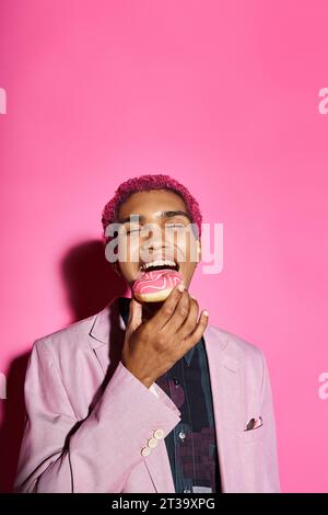 Hübscher, stilvoller Mann mit lockigen rosa Haaren in klassischem Blazer, der Donut isst, der auf rosa Hintergrund posiert Stockfoto