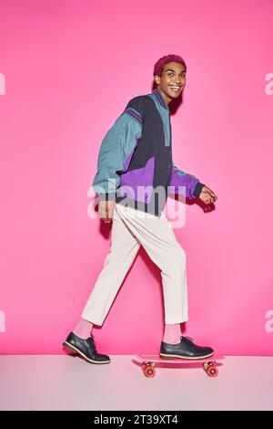 Fröhliches, junges männliches Model posiert auf Skateboard im Profil und lächelt vor rosa Hintergrund in die Kamera Stockfoto