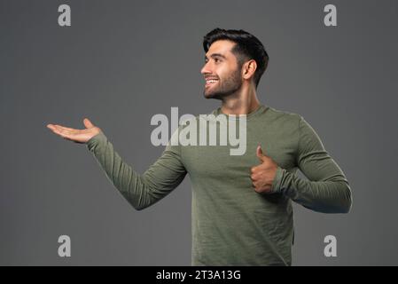 Gutaussehender, lächelnder junger kaukasischer Mann, der die Hand öffnet und seitlich schaut und in hellgrauem, isoliertem Hintergrundstudio-Foto die Schläge aufgibt Stockfoto