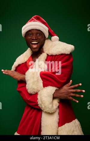 Überfröhlicher afroamerikanischer Mann im weihnachtsmannkostüm, der Gesten trägt und die Kamera auf Grün betrachtet Stockfoto