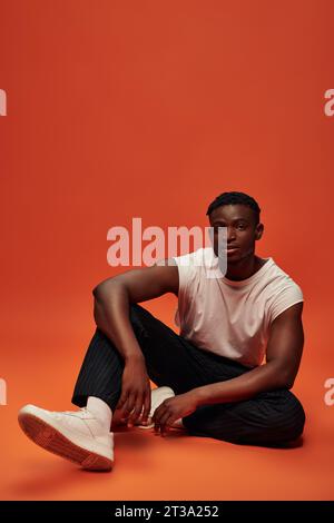 afroamerikanischer Mann in trendiger Freizeitkleidung sitzend und in die Kamera schauend vor rotem und orangenem Hintergrund Stockfoto
