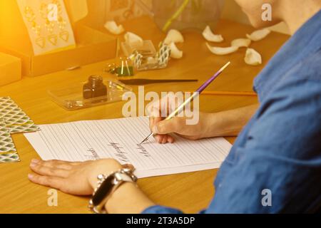 Die weibliche Hand schreibt mit dem Tintenstift die Worte danke und Kalligrafie auf ein weißes Papierblatt mit Streifen. Schreibwaren auf dem Schreibtisch aus nächster Nähe. Zauberspruch Stockfoto