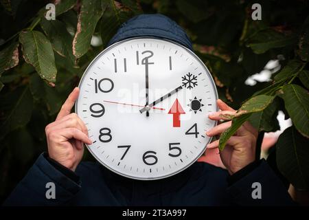 24. Oktober 2023: Mann hält große rund-um-Uhr vor seinem Gesicht mit Sommer- und Winterzeit in der Hand. Ein großer roter Pfeil steht für das Zurücksetzen der Uhr. Wechselzeiterinnerung FOTOMONTAGE *** Mann hält große runde Uhr vor seinem Gesicht mit Sommer und Winterzeit in der Hand. Ein großer roter Pfeil steht für die Uhr zurückstellen. Bild umstellen erinnerung FOTOMONTAGE Credit: Imago/Alamy Live News Stockfoto