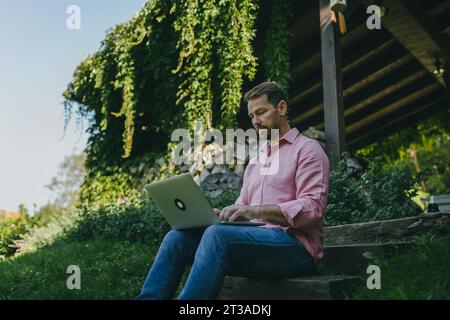 Niedrigwinkelaufnahme eines Mannes, der draußen im Garten arbeitet, mit Laptop auf den Beinen. Geschäftsmann, der aus der Ferne arbeitet und über neue Geschäfte nachdenkt Stockfoto