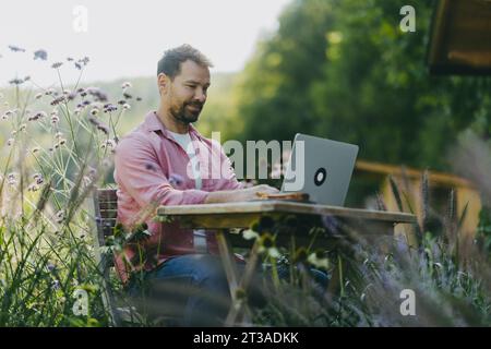 Niedrigwinkelaufnahme eines Mannes, der draußen im Garten arbeitet, mit Laptop auf den Beinen. Geschäftsmann, der aus der Ferne arbeitet und über neue Geschäfte nachdenkt Stockfoto