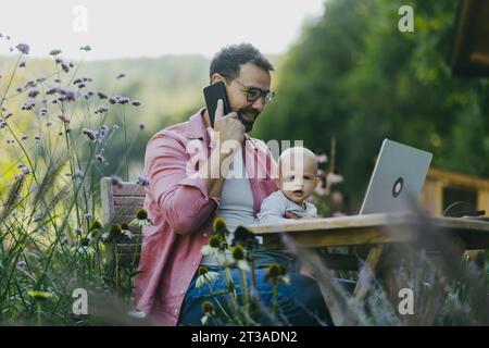 Vater hält Baby in der Hand, während er draußen im Garten an einem Laptop arbeitet. Geschäftsmann, der von der Zentrale aus arbeitet, telefoniert mit dem Kunden und Stockfoto