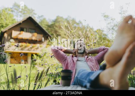 Entspannter Mann, der im Garten sitzt und die Füße auf dem Tisch liegen. Vater hat Zeit für sich, während seine Kinder in der Schule sind. Stockfoto