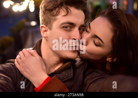 Ein junges, trendiges verliebtes Paar läuft eine Nachtstraße entlang und nimmt Selfies. Das Mädchen küsst ihren Freund. Stockfoto