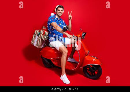 Foto in voller Länge von netten jungen männlichen präsentiert Lieferung Moped V-Zeichen tragen santa claus Druck x-MAS Kleid isoliert auf rotem Hintergrund Stockfoto