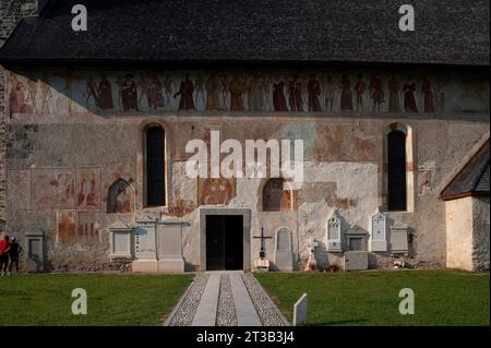 Fresken an der Südwand der Chiesa di San Vigil (Kirche St. Vigil oder Kirche St. Vigil von Trient) in Pinzolo, Trentino-Südtirol, Italien. Entlang der Mauer, unterhalb der Traufe, befindet sich ein Freskenfries Tanz des Todes, gemalt 1539 von Simone II. Baschenis (ca. 1495–1555). Unter anderen Fresken darunter befindet sich ein Teil des Heiligen Christoph und Christuskind (Mitte), ein Bischof in einer Freskennische (links unten) und ein Kreuzigungsfresko in einer anderen Nische (rechts unten). Stockfoto