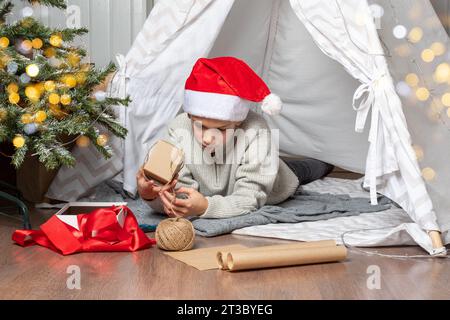 Familienweihnachtsfest. Kinder öffnen Weihnachtsgeschenke. Ein Teenager mit Weihnachtsmannmütze öffnet ein Geschenk und lächelt, während er auf dem Boden des Hauses sitzt Stockfoto