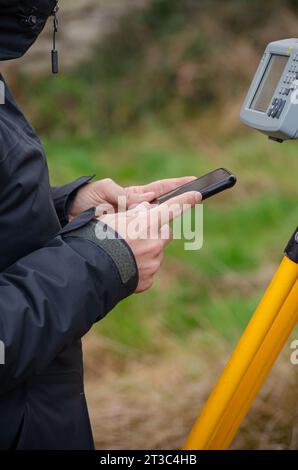 Details der Hände eines Vermessungsbeamten, die die Gesamtstation mit dem Mobiltelefon konfigurieren, topografische Arbeiten Stockfoto