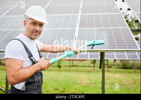 Mann, der PV-Solarpanel mit Mopp reinigt. Professioneller Arbeiter, der gute Arbeit bei der Reinigung des Solarmoduls von Staub leistet. Mann hält Mopp und lächelt direkt in die Kamera. Stockfoto