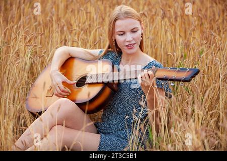 Schöne junge Frau, die im Weizenfeld sitzt und Akustikgitarre spielt Stockfoto