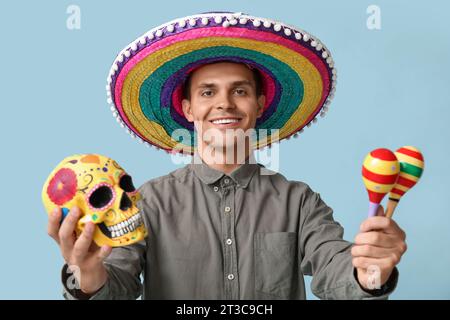 Schöner junger Mann in Sombrero mit Maracas und bemaltem Schädel auf blauem Hintergrund Stockfoto