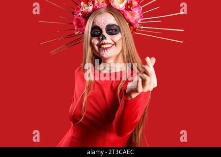 Junge Frau mit gemaltem Schädel im Gesicht, Herzgeste auf rotem Hintergrund. Feier des mexikanischen Todestages (El Dia de Muertos) Stockfoto