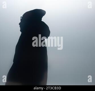 Silhouette eines Mannes, der durch ein mattiertes Fenster in Parka mit Hut und Brille gesehen wird, halblanges Profil Stockfoto