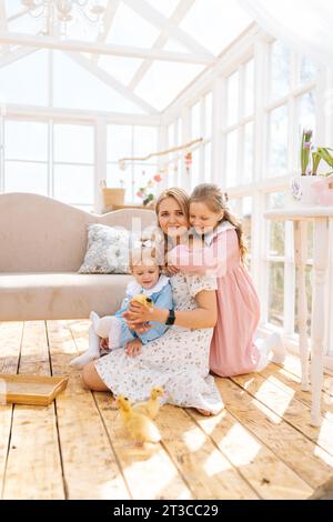 Vertikales Porträt der liebevollen blonden Mutter und zwei entzückenden Töchtern im Kleid, die mit kleinen gelben Entlein am sonnigen Tag im Haus des Sommerhauses spielen. Stockfoto