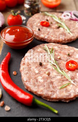 Rohe, frisch gemahlene Rinderburger, Rosmarin, Gemüse und Gewürze auf einer schwarzen Steinplatte auf Holzhintergrund. Zutaten für einen Hamburger. Stockfoto