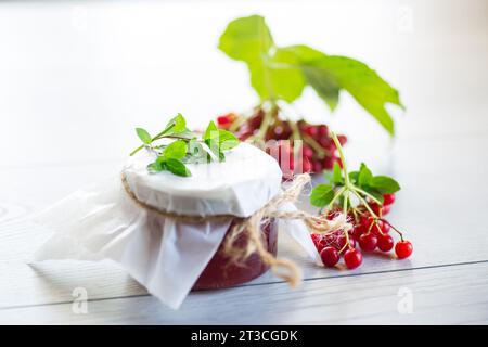 Viburnum-Marmelade in einem Glasgefäß und frisches rotes Viburnum auf einem hellen Holztisch. Stockfoto