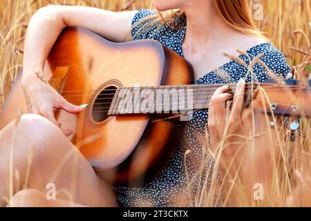 Schöne junge Frau, die im Weizenfeld sitzt und Akustikgitarre spielt. Stockfoto