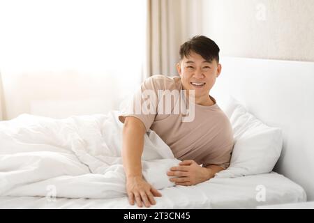 Ein glücklicher reifer asiatischer Mann wacht auf, sitzt auf dem Bett Stockfoto