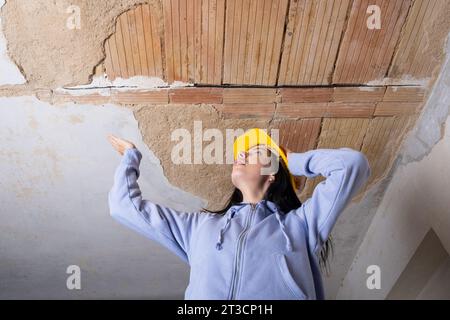 Eine junge Frau mit Schutzhelm blickt auf ein beschädigtes Loch in der Decke, wo der Putz aufgrund von Feuchtigkeitsschäden zerbröckelt ist. Sie engagiert sich in t Stockfoto
