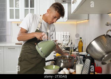 Mann, der Wasser aus dem Wasserkocher in einen Topf in einer unordentlichen Küche gießt. Viele schmutzige Geschirr und Utensilien auf der Arbeitsplatte Stockfoto