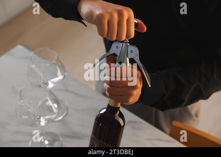 Mann öffnet eine Weinflasche mit Korkenzieher am Tisch drinnen, Nahaufnahme Stockfoto