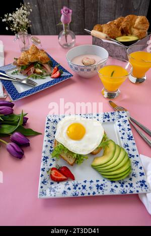 Brunch für zwei Personen mit einem wunderschönen rosafarbenen Hintergrund, Croissants, Avocados, Orangensaft, Toast-Eiern und einem parma-Sandwich. Frische Tulpen in der L Stockfoto
