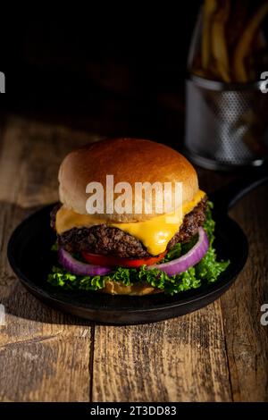 Heißer Käseeburger mit schmelzendem Käse, Salat, Tomate auf einem Brioche-Brötchen mit Pommes frites in einem konischen Halter. Dunkle Holzoberfläche Stockfoto