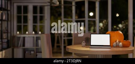 Ein runder Holztisch in einem Raum an einer Halloween-Nacht mit einem Laptop-Modell mit weißem Bildschirm, Kerzen, Kürbissen und Halloween-Ornamenten. Arbeitsplatz zu Hause auf H Stockfoto