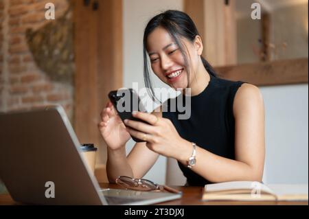 Eine fröhliche und schöne junge asiatische Frau lacht und genießt es, mit ihren Freunden zu chatten oder lustige Videos online auf ihrem Smartphone zu sehen, während sie si Stockfoto