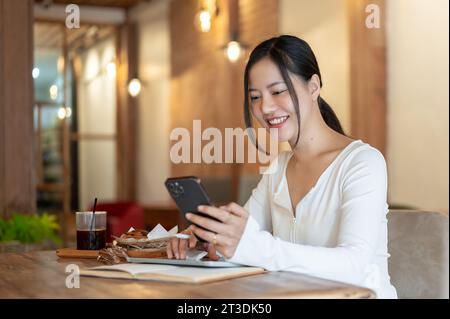 Eine glückliche und charmante junge asiatische Frau in lässiger Kleidung benutzt ihr Smartphone, schreibt ihren Freunden SMS, reagiert auf Nachrichten oder liest online einen bl Stockfoto