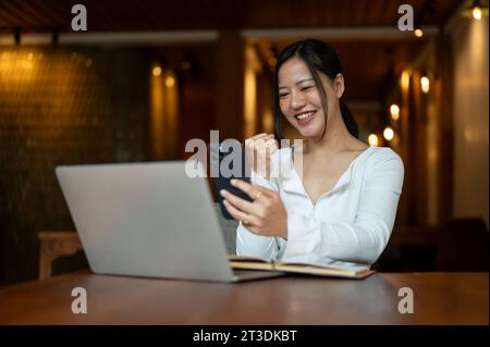 Eine fröhliche und schöne asiatische Frau schaut mit einem fröhlichen und aufgeregten Gesicht auf ihren Smartphone-Bildschirm, zeigt ihre Faust und fühlt sich überrascht von der Göttin Stockfoto