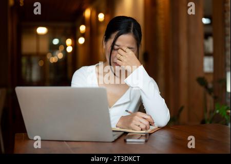 Eine gestresste und müde asiatische Frau leidet nach einem langen Arbeitstag an ihrem Projekt, der aus der Ferne in einem Café arbeitet. Stockfoto