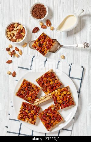 Hausgemachte Toffee gemischte Nüsse Shortbread Bars auf weißem Teller auf weißem Holztisch mit Zutaten, vertikale Ansicht von oben Stockfoto