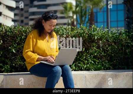 Selbstbewusste multiethnische junge Erwachsene Studentin, die online auf einem Laptop arbeitet und auf einer Steinbank vor dem Hintergrund städtischer Gebäude sitzt. Menschen. Int Stockfoto