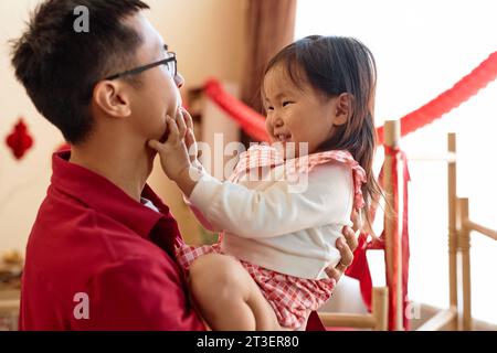 Seitenansicht Porträt eines süßen asiatischen Mädchens, das mit seinem Vater zu Hause spielt und fröhlich kichert, beide tragen rot Stockfoto