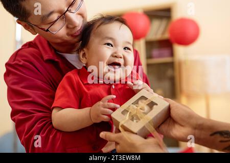 Porträt eines süßen asiatischen Babys, das eine Schachtel mit Glückskeksen hält, mit der Familie, die gemeinsam das chinesische Neujahr zu Hause feiert Stockfoto