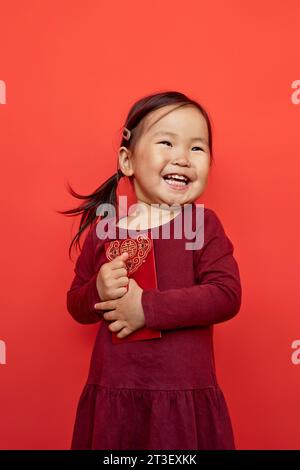Vertikales Porträt eines glücklichen asiatischen Mädchens, das hongbao-Umschlag hält und mit aufrichtiger Emotion lächelt und vor rotem Hintergrund steht Stockfoto