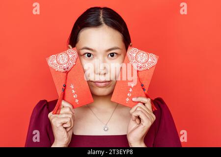 Porträt einer jungen Frau, die hongbao-Umschläge gegen Rot hält und in die Kamera blickt, ist jedes Jahr überflutet Stockfoto