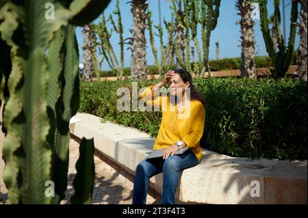 Schöne Multi-ethnische lockige junge Erwachsene nachdenkliche Frau in lässiger Kleidung, sitzt auf einer Steinbank mit einem Laptop auf den Knien, traumhafte Looki Stockfoto