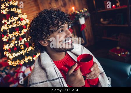 Foto träumender Good Guy mit Locken bedeckt weiße Decke Genießen Sie am Abend magische Atmosphäre Drinks Tasse Kaffee im Innenbereich Beleuchtung auf Hintergrund Stockfoto