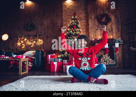 Foto in voller Länge eines jungen, flippigen arabischen Mannes, der Fäuste auf dem Sitzboden hebt und zu Hause in der Nähe des Kamins feiert Stockfoto