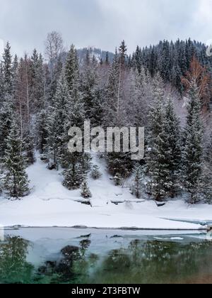 Winterlandschaft mit Bergen und einem ungefrorenen Fluss mit Reflexion. Bewölkter, schneebedeckter Tag. Sanfte Beleuchtung Stockfoto