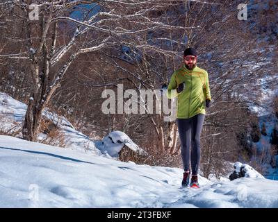 Ein bärtiger Mann joggt im Winter draußen hoch in den Bergen. Ein Kerl in grüner Jacke und Sonnenbrille trainiert. Trailrunning Stockfoto