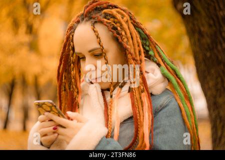 Schöne junge Frau mit Dreadlocks mit Smartphone im Herbstpark. Stockfoto